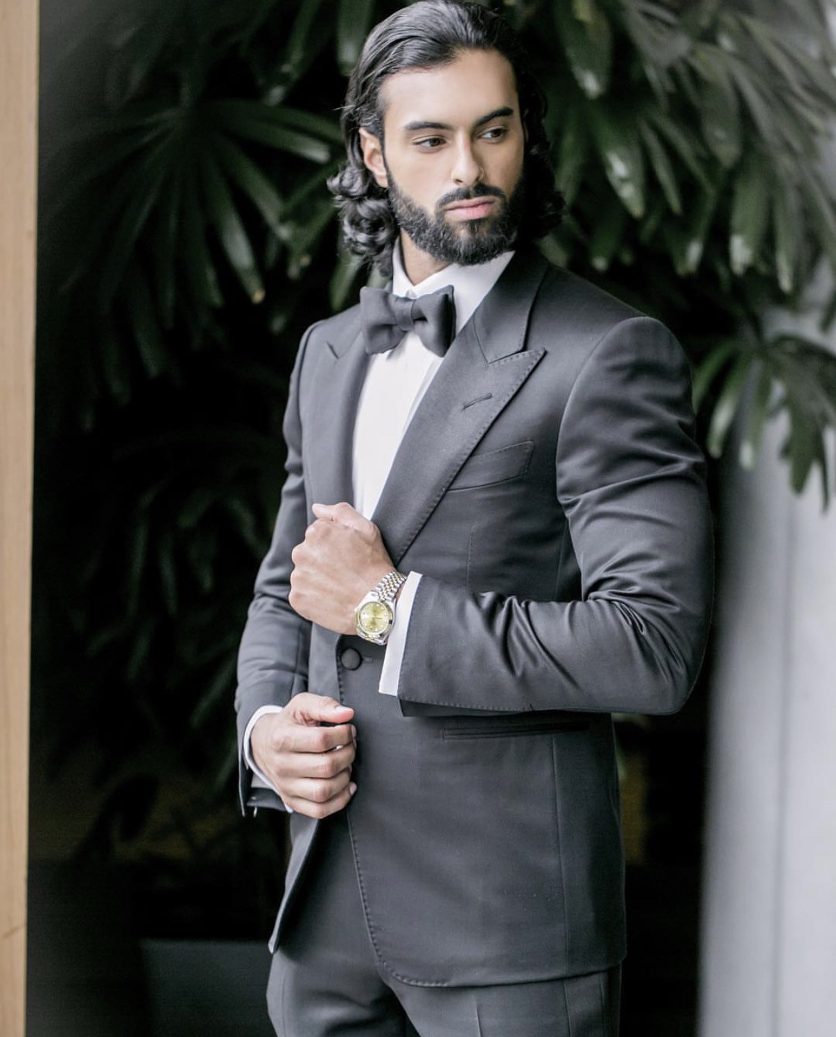 Harry Johal in a black peak-lapel tuxedo adjusting his cuff beside dark foliage.