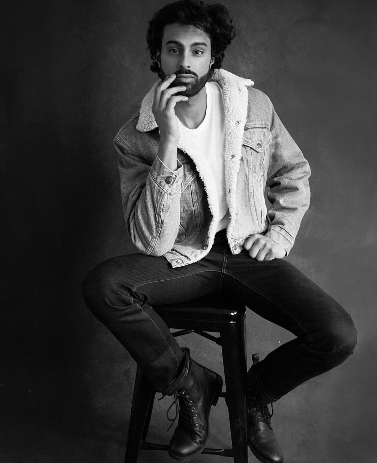 Black and white full-length portrait of Harry Johal seated on a stool in a shearling-lined denim jacket and boots.