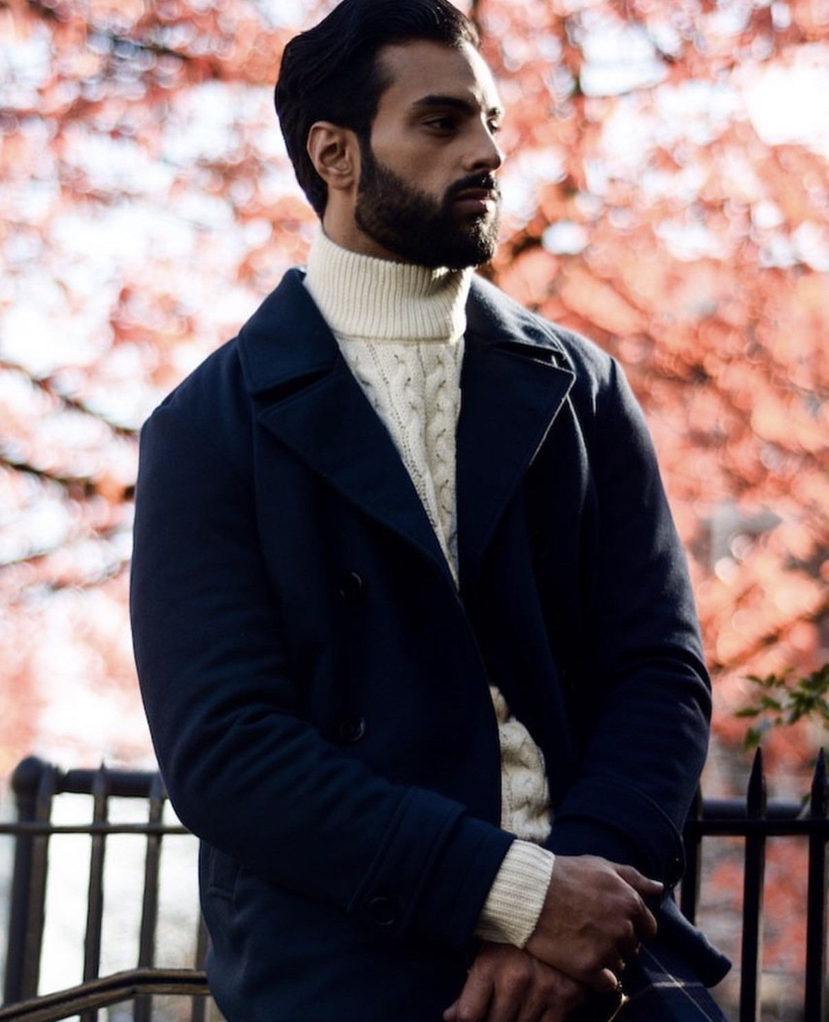 Harry Johal in a navy double-breasted overcoat and cream cable knit, autumn leaves behind him.