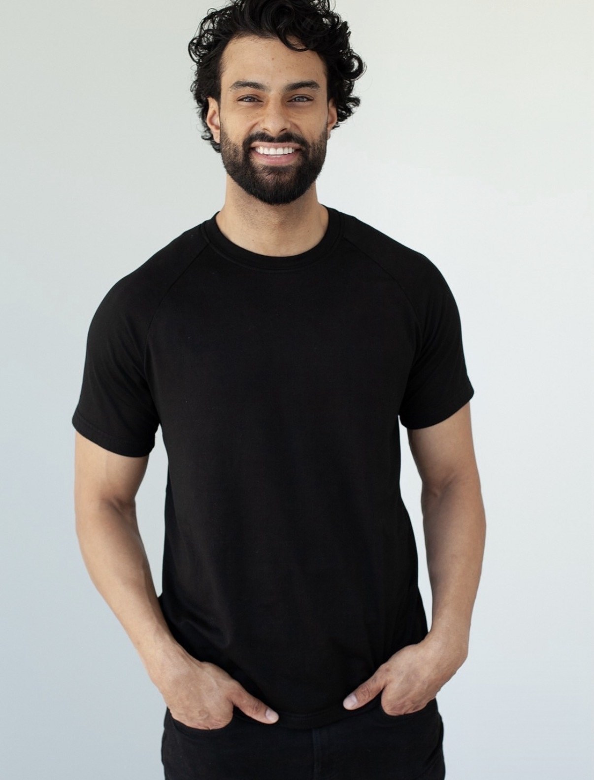 Harry Johal smiling in a black crew-neck t-shirt against a plain light studio background.