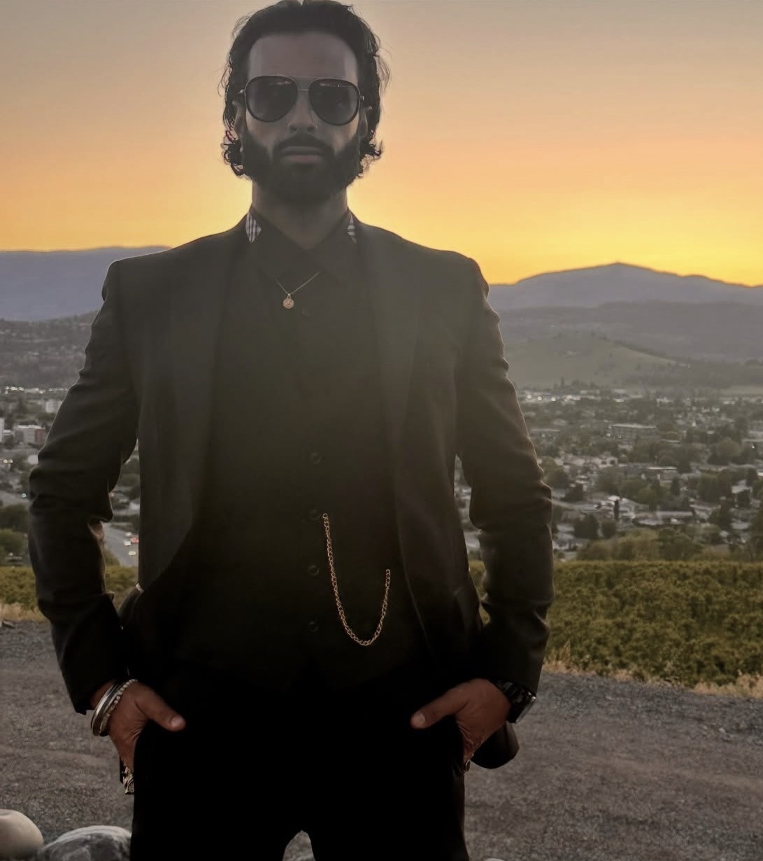 Harry Johal in a black suit and sunglasses at sunset above the city.